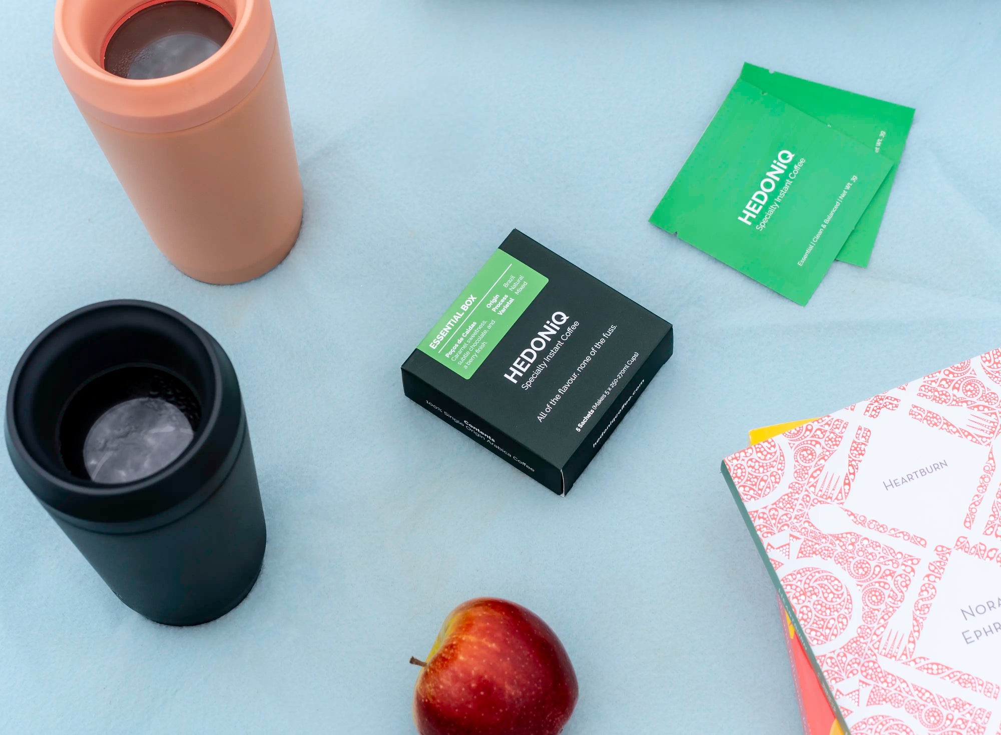 Hedoniq Essential Box with sachets, reusable TOPL travel cups, an apple and books on a picnic blanket, perfect for coffee anywhere, anytime.