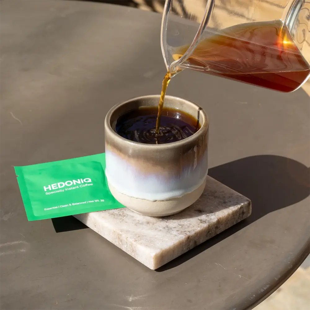 Pouring Hedoniq specialty instant coffee into a ceramic cup, with a green Essential Box sachet on a marble coaster in natural light.