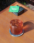 Glass of iced Hedoniq specialty instant coffee on a wooden outdoor table with Essential Box sachet and packaging.