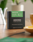 Hedoniq Essential Box specialty instant coffee on a wooden table with a yellow coffee cup and gooseneck kettle in the background.