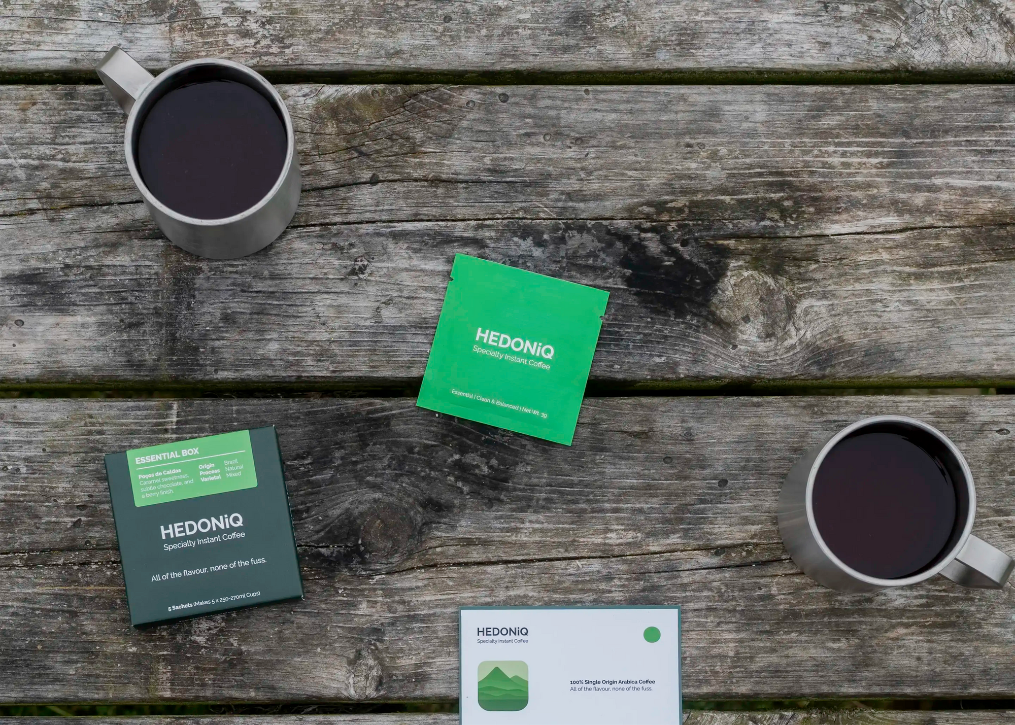 Hedoniq Essential Box specialty instant coffee with sachet, postcard, and two cups of brewed coffee on a rustic wooden bench.