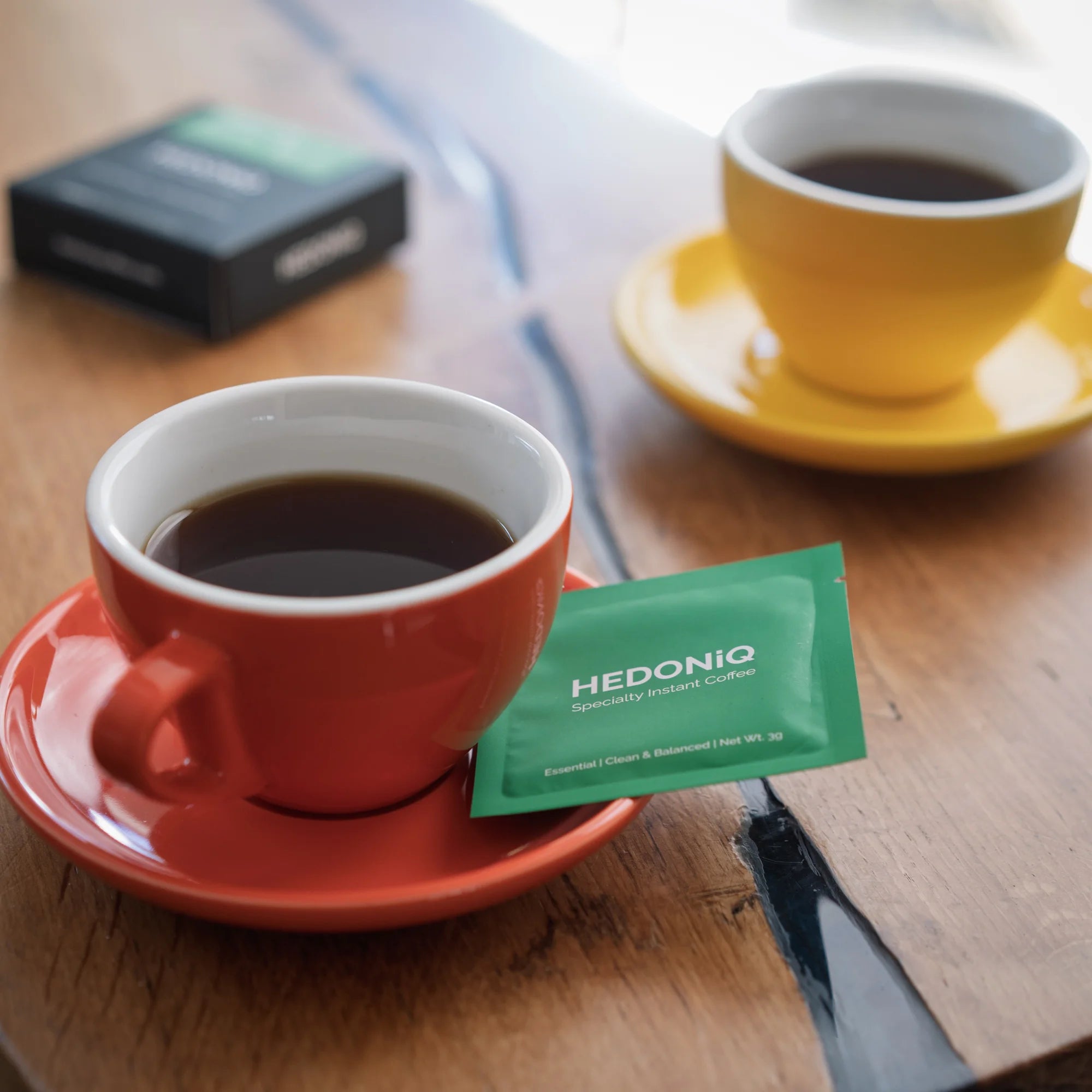 Two colourful cups of brewed Hedoniq specialty instant coffee with Essential Box sachet on a wooden table.