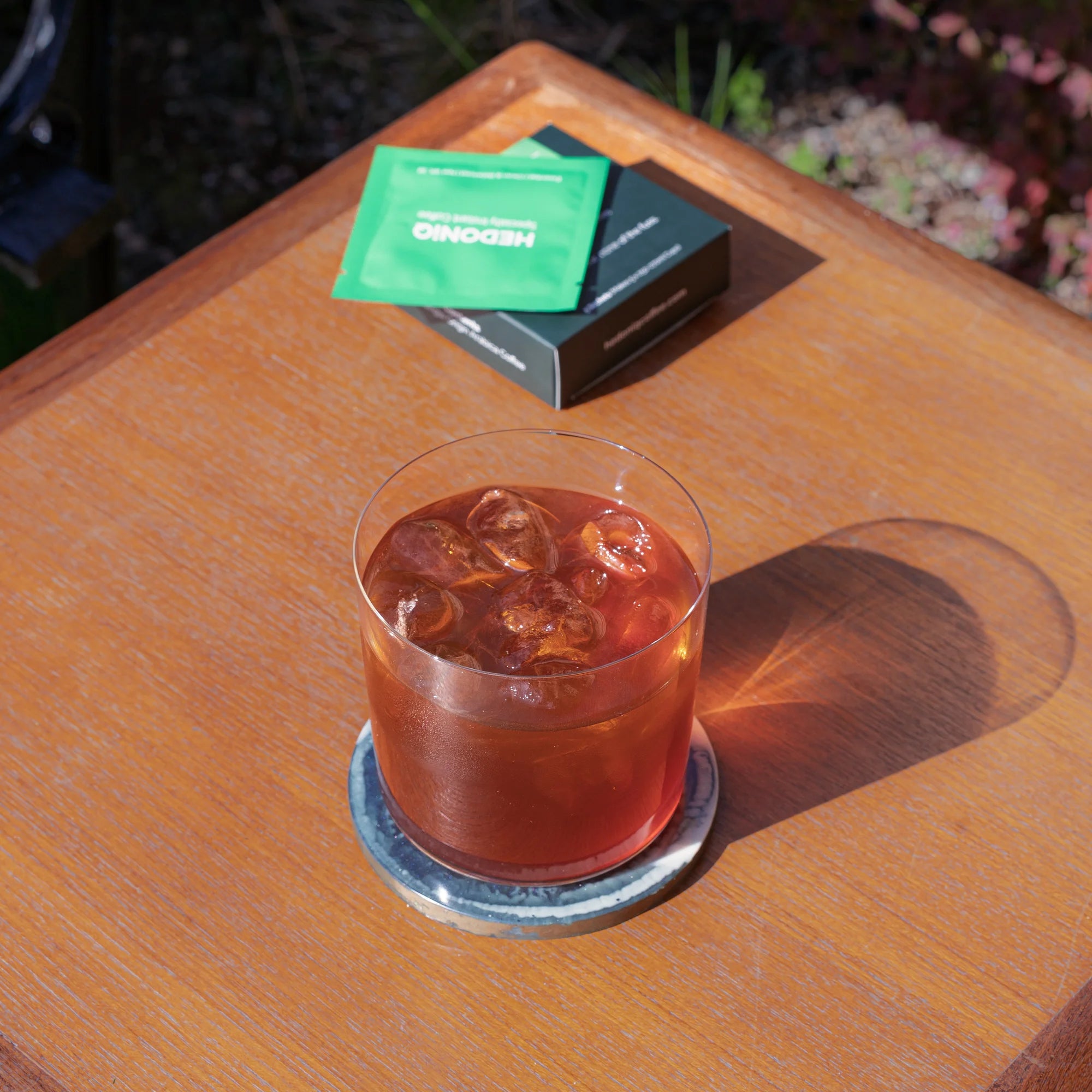 Glass of iced Hedoniq specialty instant coffee on a wooden outdoor table with Essential Box sachet and packaging.