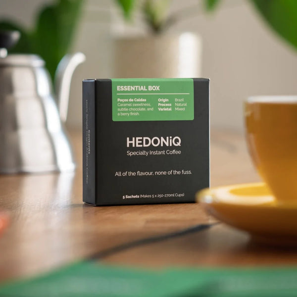 Hedoniq Essential Box specialty instant coffee on a wooden table with a yellow coffee cup and gooseneck kettle in the background.